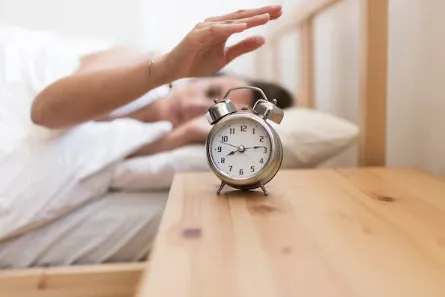 Hand reaching to stop a ringing alarm clock on a bedside table.webp
