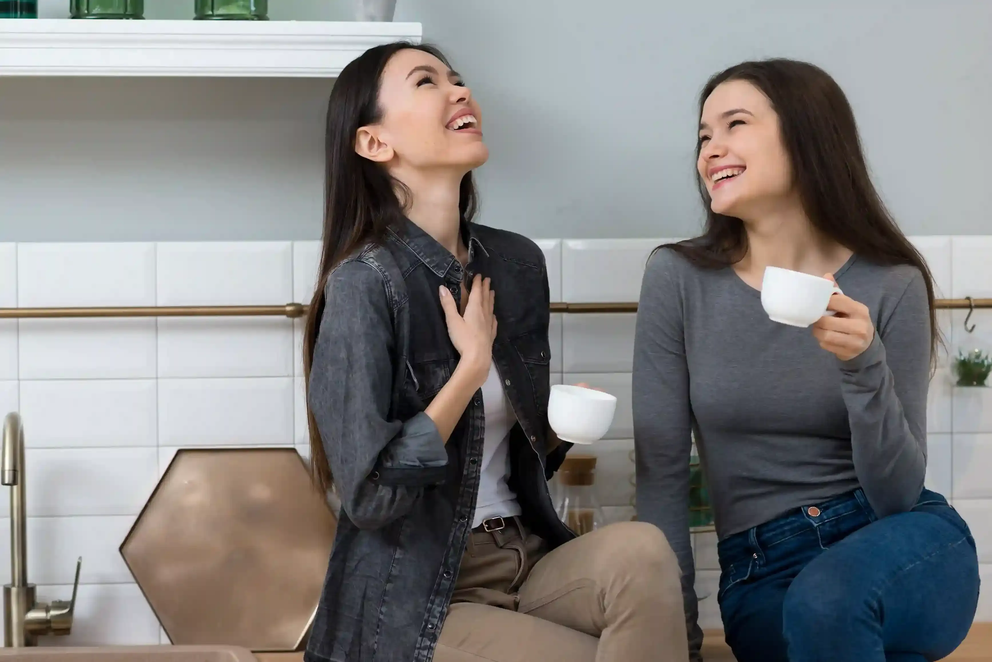 Two young women laughing and enjoying a friendly conversation in English while drinking coffee in a modern kitchen
