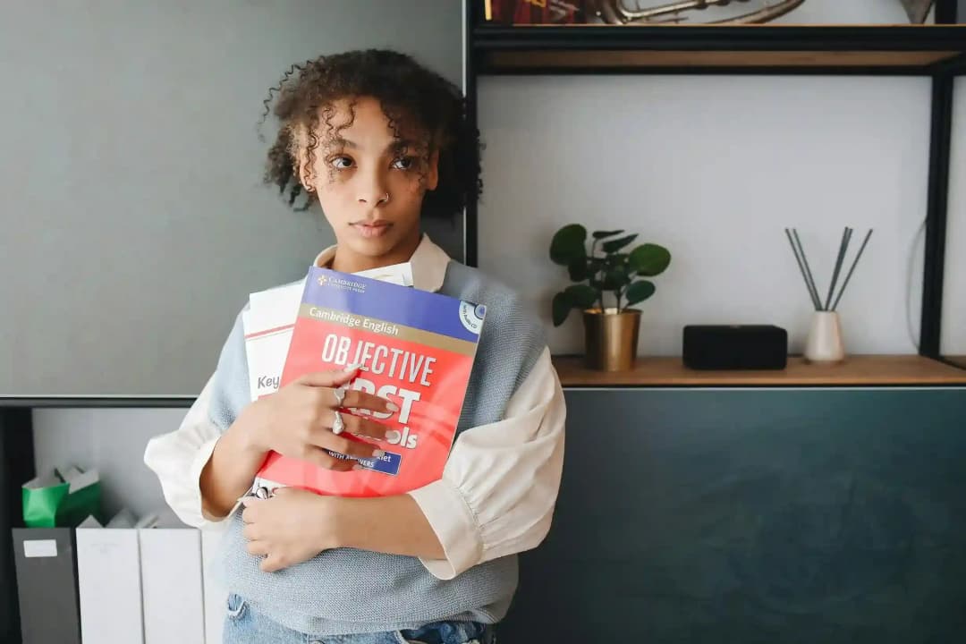 A young woman with curly hair holding Cambridge English Objective First textbooks in a modern, minimalist study area.