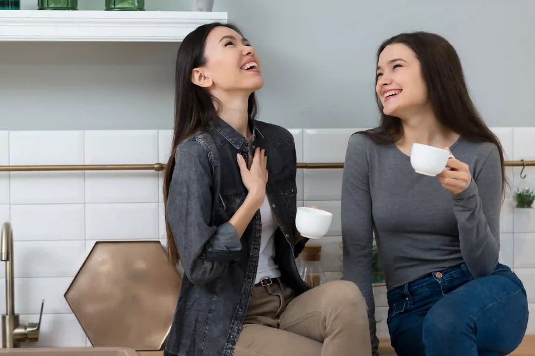 Two young women laughing and enjoying a friendly conversation in English while drinking coffee in a modern kitchen